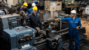 A group of four factory workers wearing blue coveralls and protective helmets operate and inspect large industrial machinery in a workshop. One worker points while holding a clipboard, directing another who adjusts a machine—underscoring the theme "From Ignored Repairs to Catastrophes: The True Cost of Neglecting Maintenance.